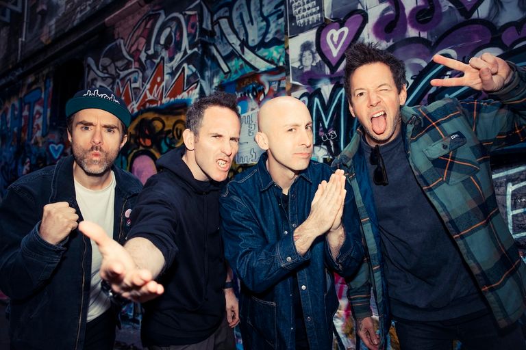 Pop-punk band Simple Plan poses together playfully with expressive faces in front of a colorful, graffiti-covered wall.